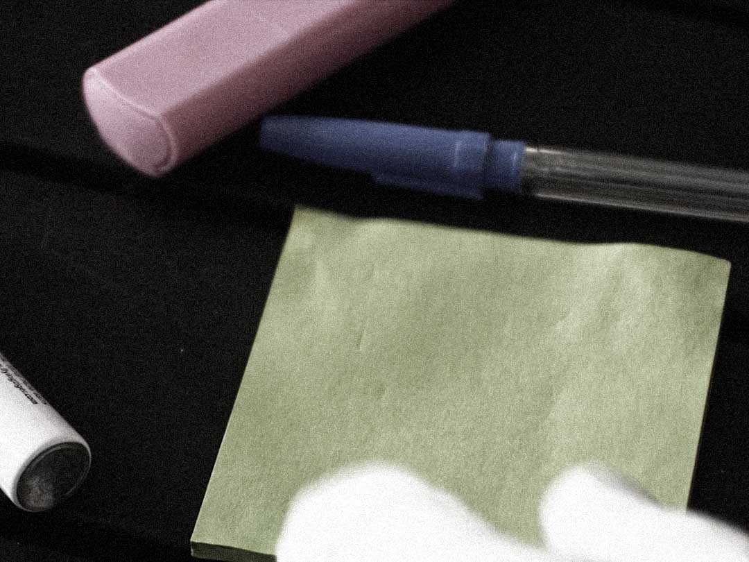 A pen, an eraser and a blank green sticky note on a dark desk.